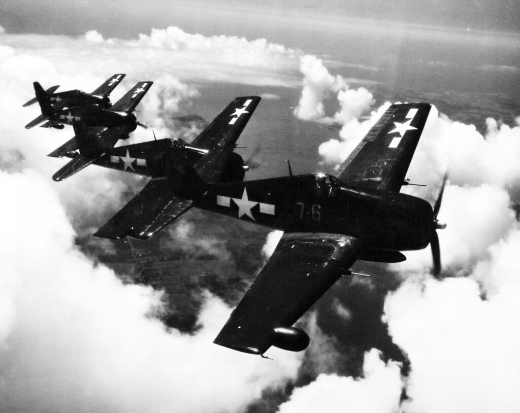 Four Grumman F6F-5N Hellcat night fighters fly in formation during the daytime over clouds. The planes are painted in a dark color to blend in with the night sky. Each has a radar pod installed on the starboard wing.