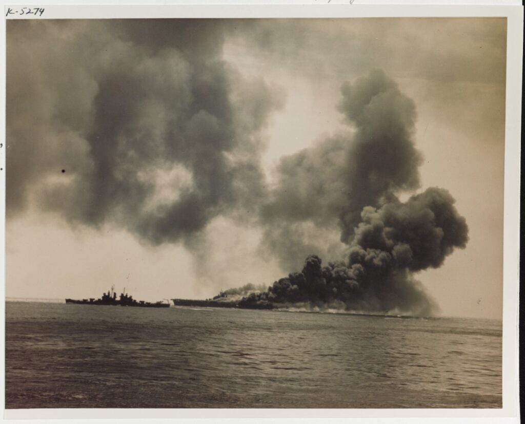 Huge pillars of smoke rise from USS Bunker Hill after two Kamikazes strike her. The photo is taken from a distance with a Cleveland-class cruiser nearby.