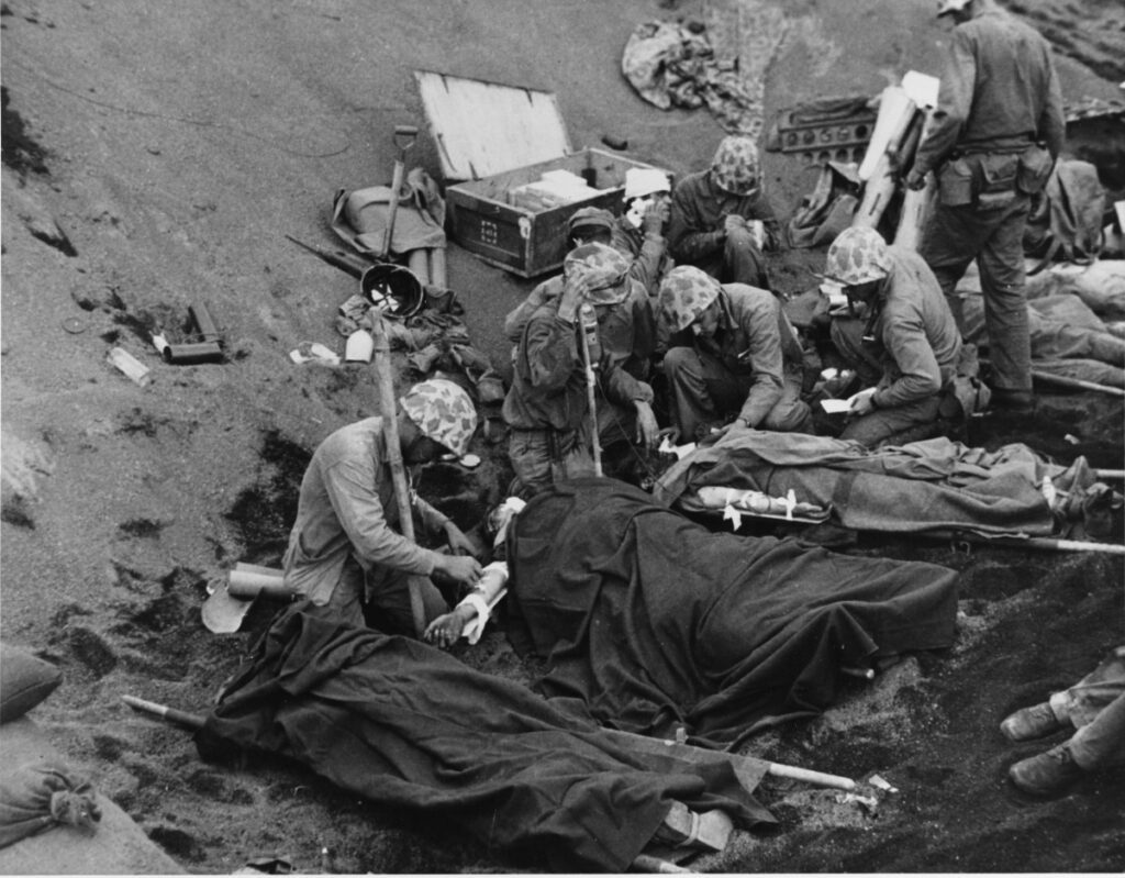 Five stretchers with wounded marines lie side by side at the bottom of a steep hill. Half a dozen medical personnel tend to their wounds while another wounded marine sits on the ground,his head wrapped in bandages. 20 February 1945, on Iwo Jima. National Archives, 80-G-435702