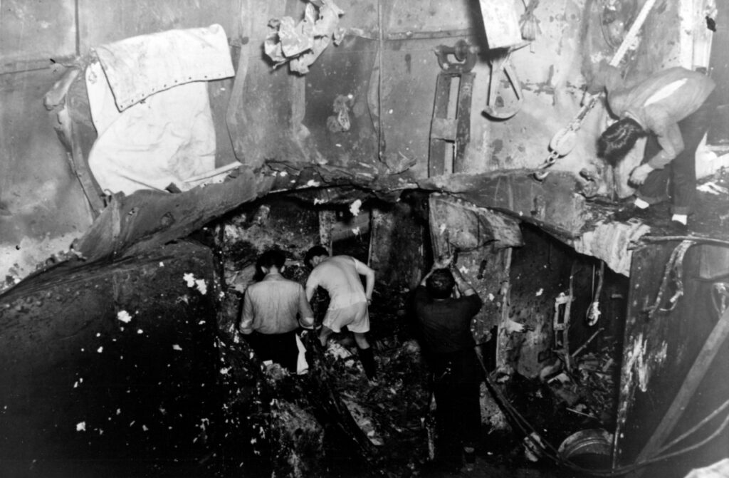 The image is of the severely damaged third and fourth decks of the USS Yorktown (CV-5) after a 250 pound bomb detonated on those decks during the Battle of the Coral Sea. There is a hole in the third deck through which can be seen two men working amongst the twisted metal. A third man stands hear the edge of the photo looking down from the dege of the hole.