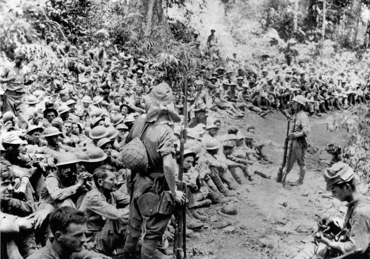 Taken just before the Bataan Death March starts, more than 100 US soldiers are shown sitting down while being guarded by several Japanese soldiers carrying rifles with bayonets. A Japanese photographer is shown on the right adding film to his camera.