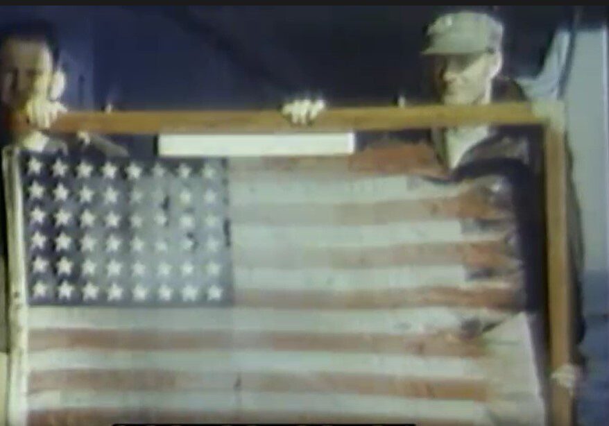 Two officers hold the USS Franklin's flag in April 1945. The flag, which flew from the island during the 19 March attack, is encased in glass within a wooden frame. (Screenshot from a US Navy film.)